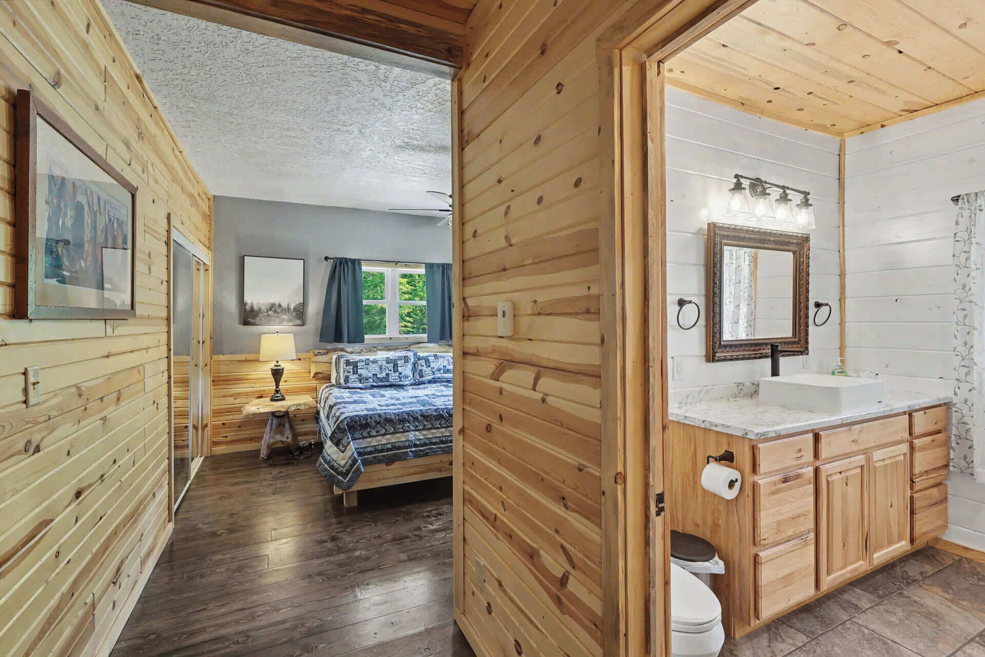 A wooden cabin interior with a bed, a bathroom with a sink and mirror, and a window with curtains.