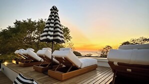 En terrasse med liggestole og en stribet parasol, med udsigt til en solnedgang.