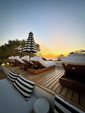 A deck with lounge chairs and a striped umbrella, overlooking a sunset. - NOAM by the sea Ocean View Suite #1 Adults Only (Tamarindo)
