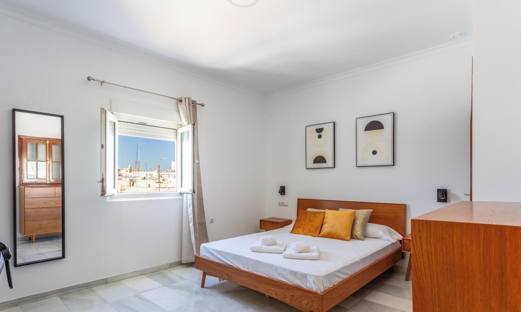A hotel room with a bed, a nightstand, a wooden dresser, a window with a view of buildings, and two framed artworks on the wall.