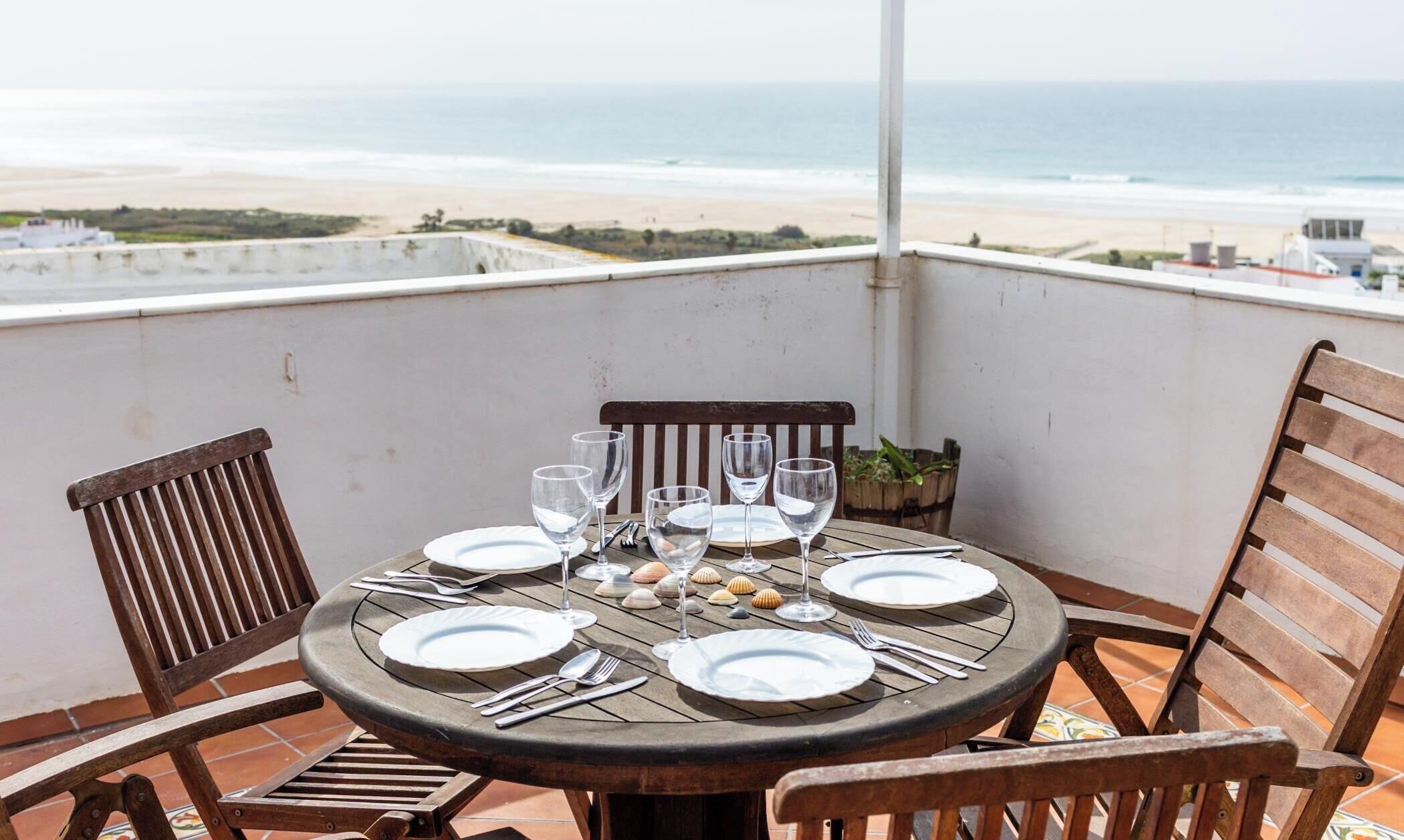 Une table ronde dressée pour quatre personnes, avec assiettes, couverts et verres à vin, donnant sur une plage.