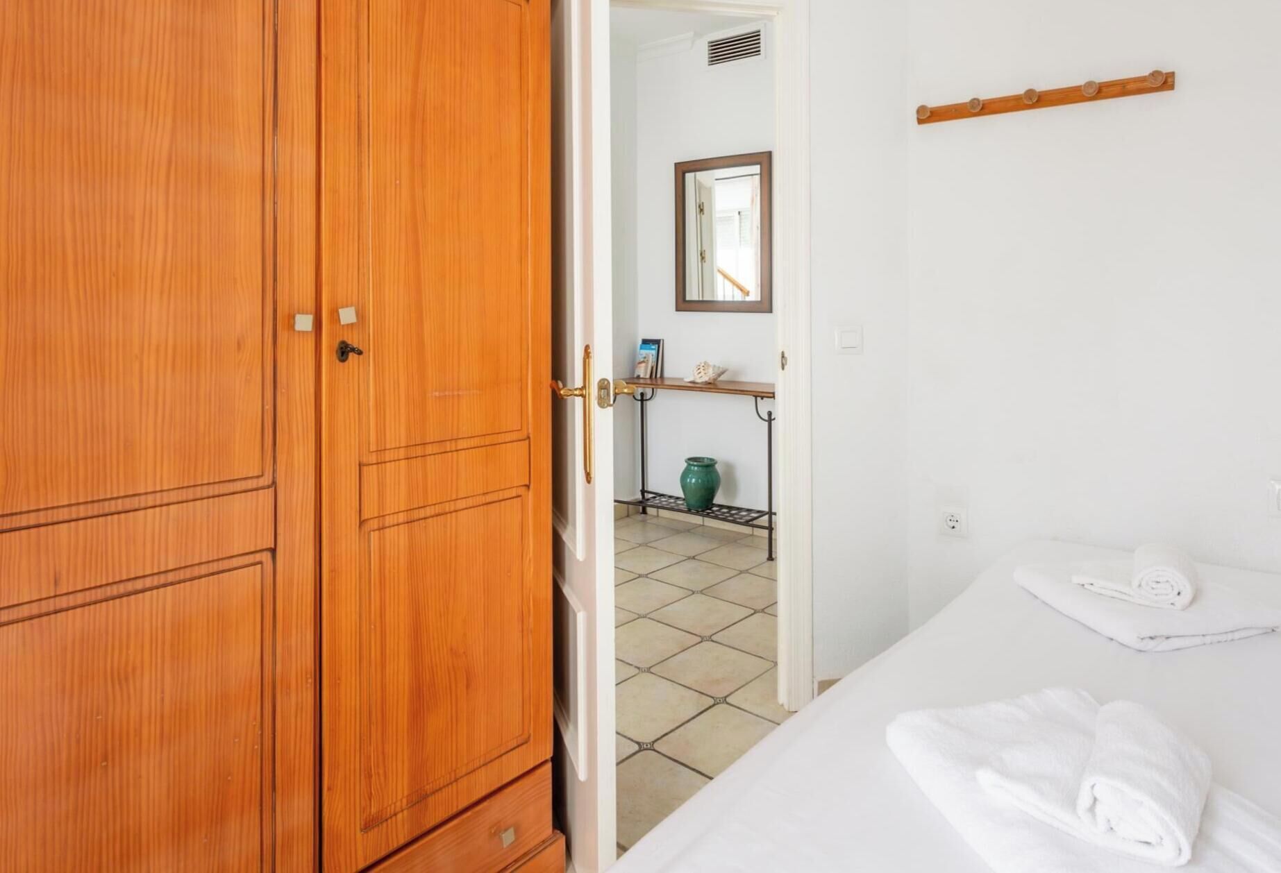 Une pièce avec une armoire en bois, un lit recouvert d’une couette blanche, une petite étagère avec un vase et un miroir accroché au mur.