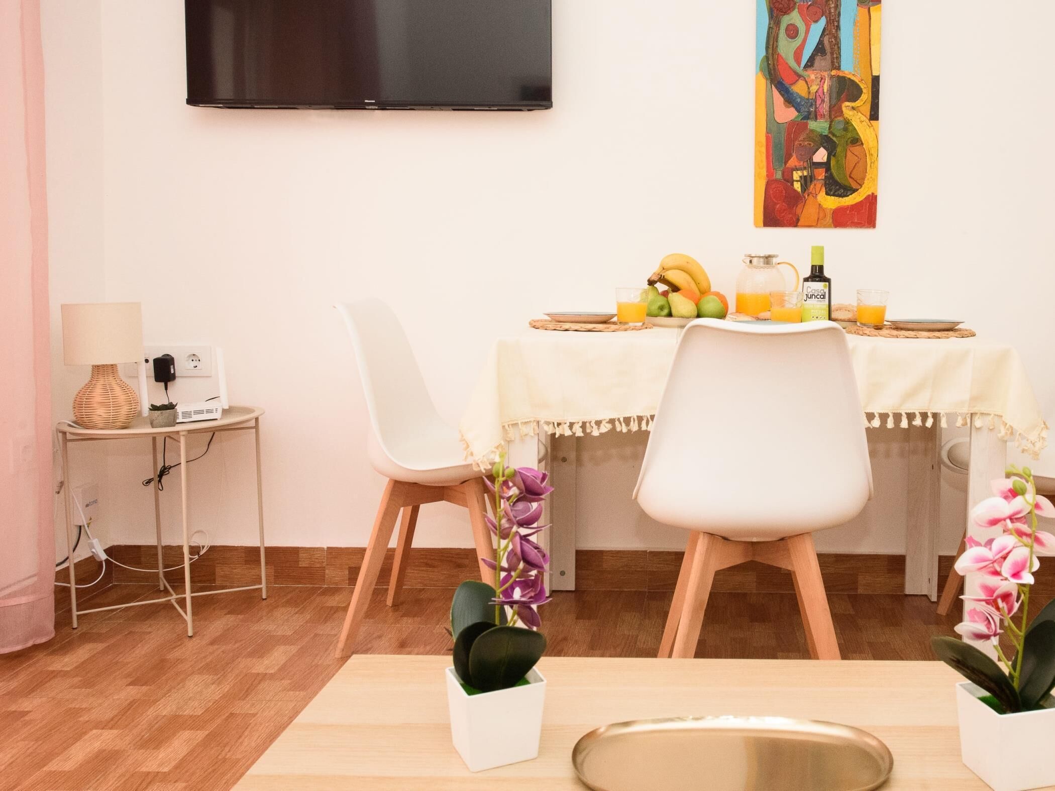 A dining area with a table set for two, a TV mounted on the wall, and a small side table with a lamp and power outlet.