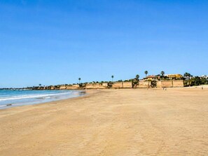 A wide sandy beach with a clear blue sky, a few palm trees, and a building with a red roof in the background. - Luz del Sur El Puerto by Apartments Time (El Puerto de Santa María)