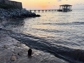 Eine Küstenlandschaft mit einer hölzernen Pier, die sich über ruhiges Wasser erstreckt, einer felsigen Uferlinie und einem gazeboartigen Bauwerk am Ende der Pier.