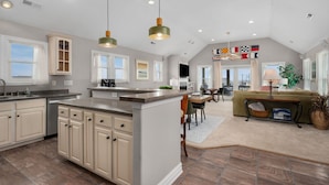 A spacious kitchen with a large island, a dining area with a table and chairs, and a living room with a sofa and a TV. - Kitty Hawk Vacation Home with Pool, Sleeps 10 (Kitty Hawk)
