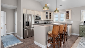 A kitchen with a central island, stainless steel appliances, and wicker bar stools. - Kitty Hawk Vacation Home with Pool, Sleeps 10 (Kitty Hawk)
