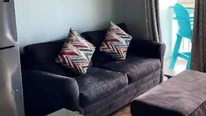 A living room with a sofa, two patterned cushions, a small table, and a window with curtains. - Front Row Beachfront Bliss Awaits! Panoramic View! (Galveston)