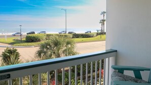 A balcony with a view of a parking lot, a beach, and palm trees. - Front Row Beachfront Bliss Awaits! Panoramic View! (Galveston)
