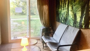 A room with a chair, a table with a lamp, a window with a view of greenery, and a forest-themed wall.