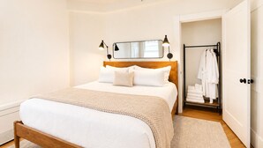 Ein Schlafzimmer mit einem hölzernen Bett, weißer Bettwäsche, einem Spiegel über dem Bett und einem begehbaren Kleiderschrank mit aufgehängten Bademänteln.