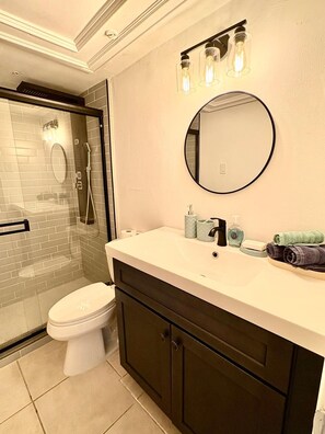 Un baño con ducha envuelta en cristal, inodoro, tocador con espejo y artículos de aseo.