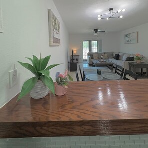 A dining area with a wooden table, two potted plants, and a view into a living room with a sofa and a ceiling fan. - Cozy 2BR Condo in Houston Medical Center, Galleria, downtown Sleeps 4 (Houston)