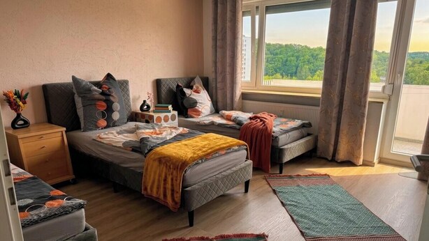 A bedroom with two beds, a window with curtains, a bedside table with a vase, and a view of greenery outside. - Stylish City Retreat – Your Temporary Home in Würzburg for 7 People (Würzburg)