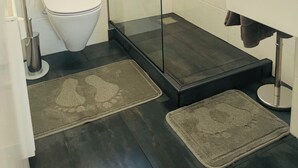 A bathroom with a glass shower enclosure, a white toilet, a sink with a towel, and two rugs on the floor. - Stylish City Retreat – Your Temporary Home in Würzburg for 7 People (Würzburg)