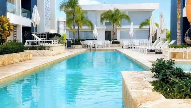 Ein Swimmingpool mit Liegestühlen und Sonnenschirmen, umgeben von Palmen und modernen Gebäuden.