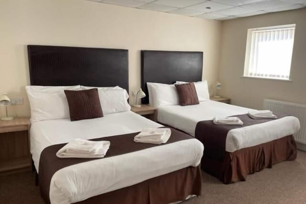 A hotel room with two beds, each with a headboard, white linens, and brown accents. There is a window with blinds, a nightstand with a lamp, and a radiator.