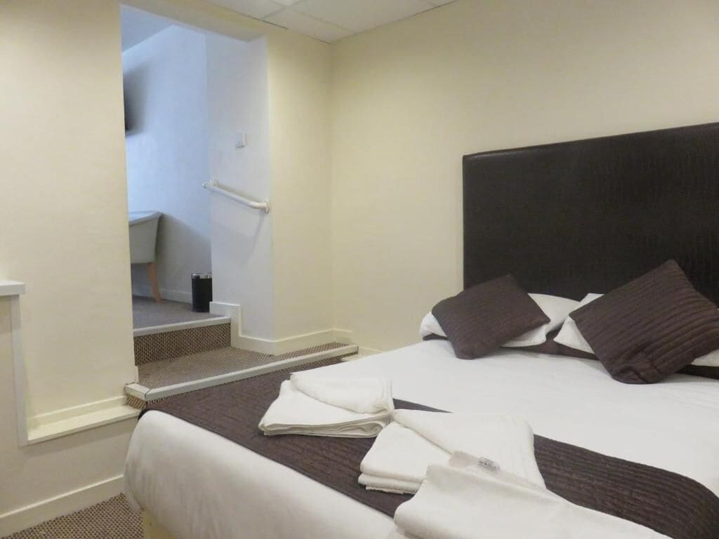 A hotel room with a bed, pillows, a bedside table, and a view of a staircase.