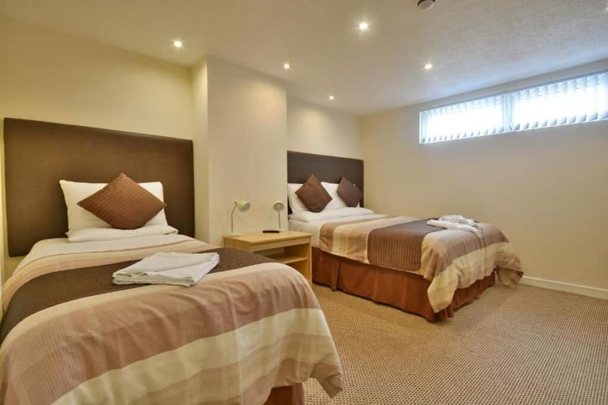 A hotel room with two beds, a nightstand, a lamp, and a window with blinds.