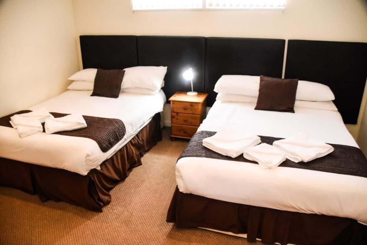 Two twin beds with white bedding and brown headboards, a nightstand with a lamp, and a window with blinds.