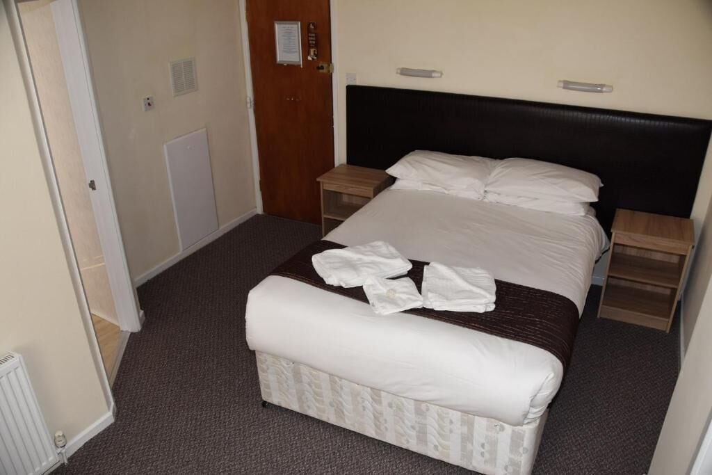 A hotel room with a bed, a bedside table, a nightstand, a bench, and a door.