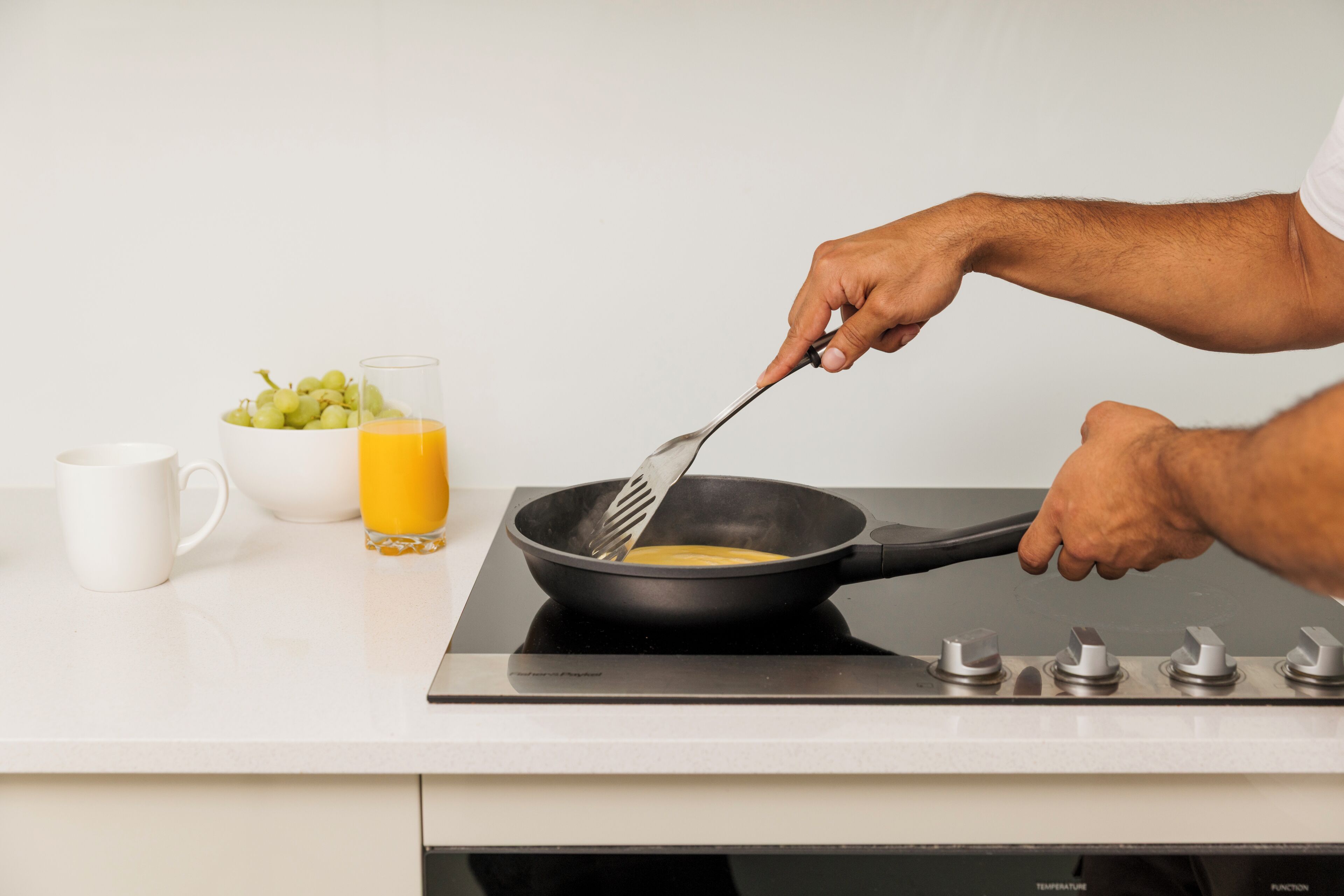 Una cocina con cubierta blanca, un bol de uvas, un vaso de zumo de naranja y una sartén sobre una cocina de inducción.
