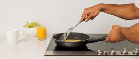 Una cocina con cubierta blanca, un bol de uvas, un vaso de zumo de naranja y una sartén sobre una cocina de inducción.
