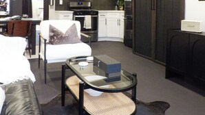 Una sala moderna con sofá, una mesita de centro de vidrio, un rincón de comedor con una silla y una cocina equipada.