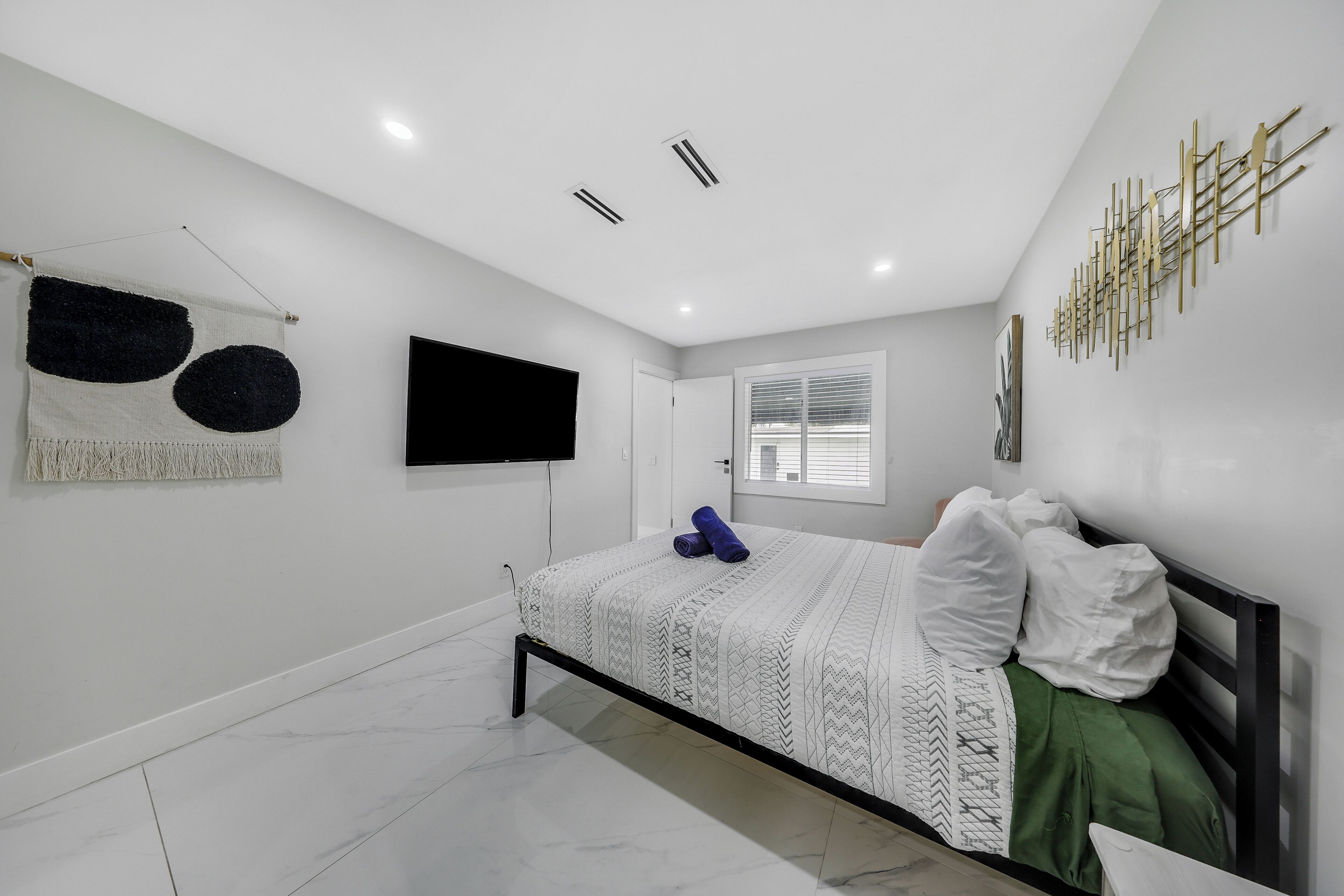 A modern bedroom with a bed, a flat-screen TV, a wall-mounted artwork, and a window with blinds.