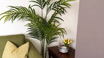 A room with a green sofa, a large potted plant, a small wooden table with a vase of yellow flowers, and a decorative item.