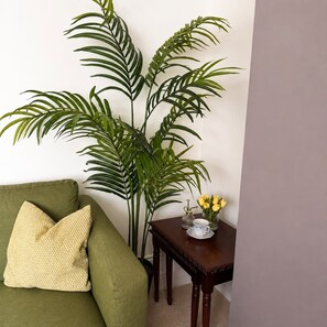 Ein Zimmer mit einem grünen Sofa, einer großen Topfpflanze, einem kleinen Holztisch mit einer Vase voller gelber Blumen und einem dekorativen Gegenstand.