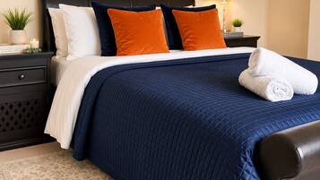 A neatly made bed with a dark blue quilt, white pillows, and two orange accent pillows. There is a white towel on the bed, a nightstand with a lamp, and a small plant on the nightstand.