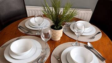 A round dining table with white plates, cutlery, and glassware, surrounded by woven placemats and a potted plant.