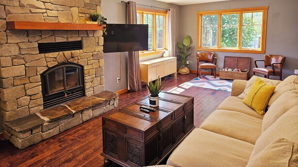 A living room with a stone fireplace, a flat-screen TV mounted above it, a wooden coffee table, a beige sofa with yellow pillows, and a large window with a view of greenery.