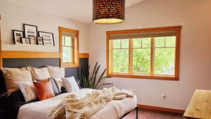 A bedroom with a bed, a window, a plant, and framed pictures on the wall.
