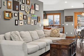 A living room with a sectional sofa, a dining table with chairs, a chandelier, and a wall decorated with framed pictures and a deer head. - Fantastic In Town Location (Fernie)