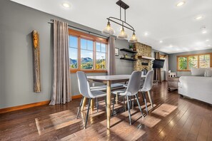 A dining area with a table and chairs, a stone fireplace, a television, and a sofa in a room with wooden flooring and large windows. - Fantastic In Town Location (Fernie)