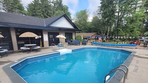 A rectangular swimming pool with a blue interior, surrounded by a concrete deck with tactile paving. There are lounge chairs and umbrellas, and a building with a dark roof and beige walls in the background. - Maple Cabin at Pine Ridge Park (Pembroke)