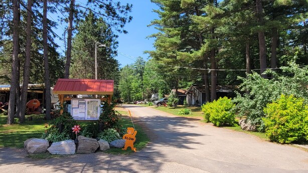 A rural road with a wooden signpost, a small wooden structure with a red roof, and dense greenery. - Maple Cabin at Pine Ridge Park (Pembroke)