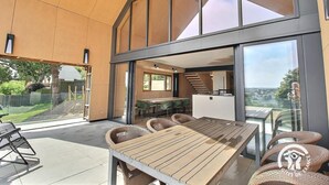 Ein modernes Haus mit einer großen Terrasse, einem Essbereich mit Korbmöbeln und Blick auf die Landschaft.