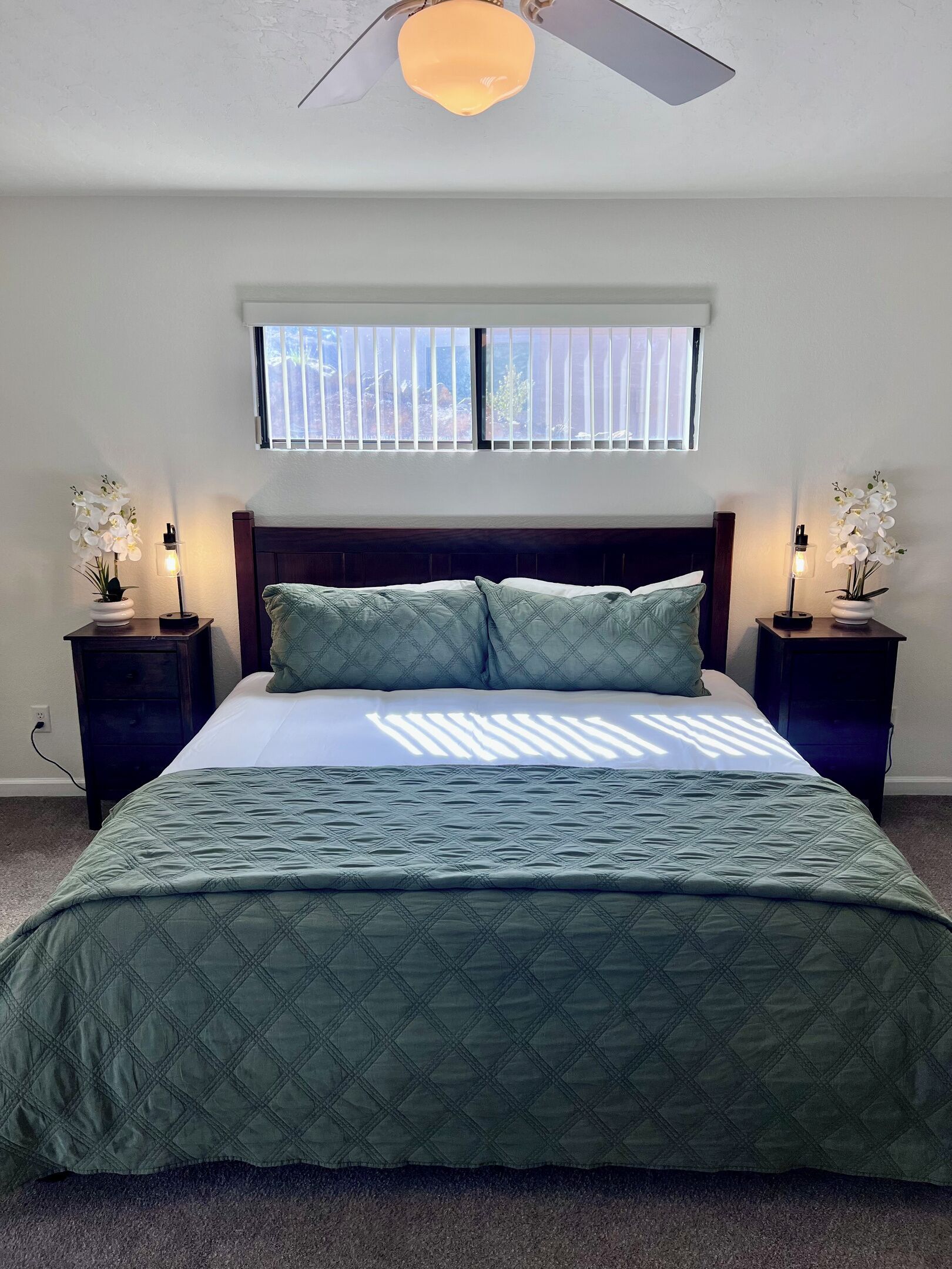 Une chambre à coucher avec un lit, deux tables de chevet avec des lampes, une fenêtre avec des stores et un ventilateur de plafond.