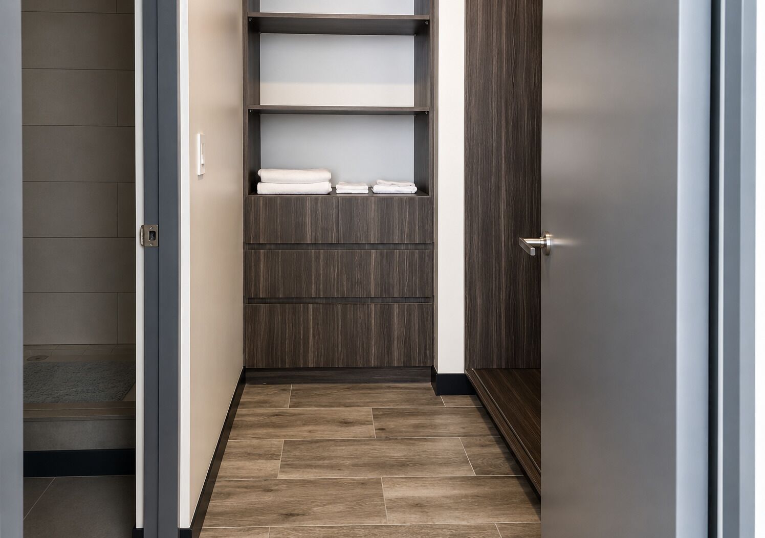A narrow hallway with a wooden floor, a built-in shelf unit with folded towels, and a door with a metallic handle.
