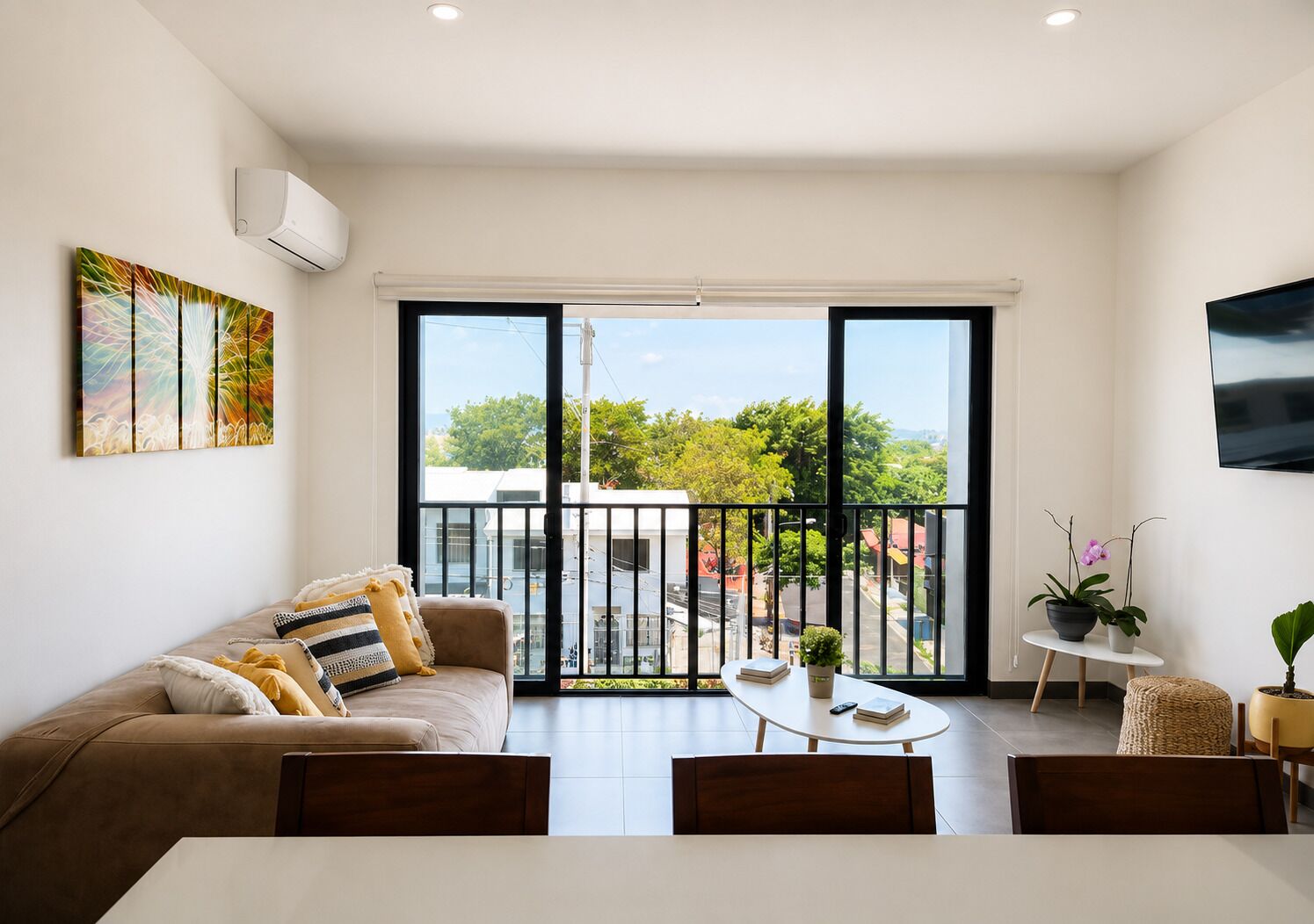 A modern living room with a sofa, a dining table, and chairs, a balcony with a view, and a mounted TV.