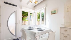 A dining area with a table set for four, white chairs, a white cabinet, and a door with a glass panel. - Le Patio de la Mer – Comfortable one-bedroom flat with terrace in Valras-Plage (Valras-Plage)