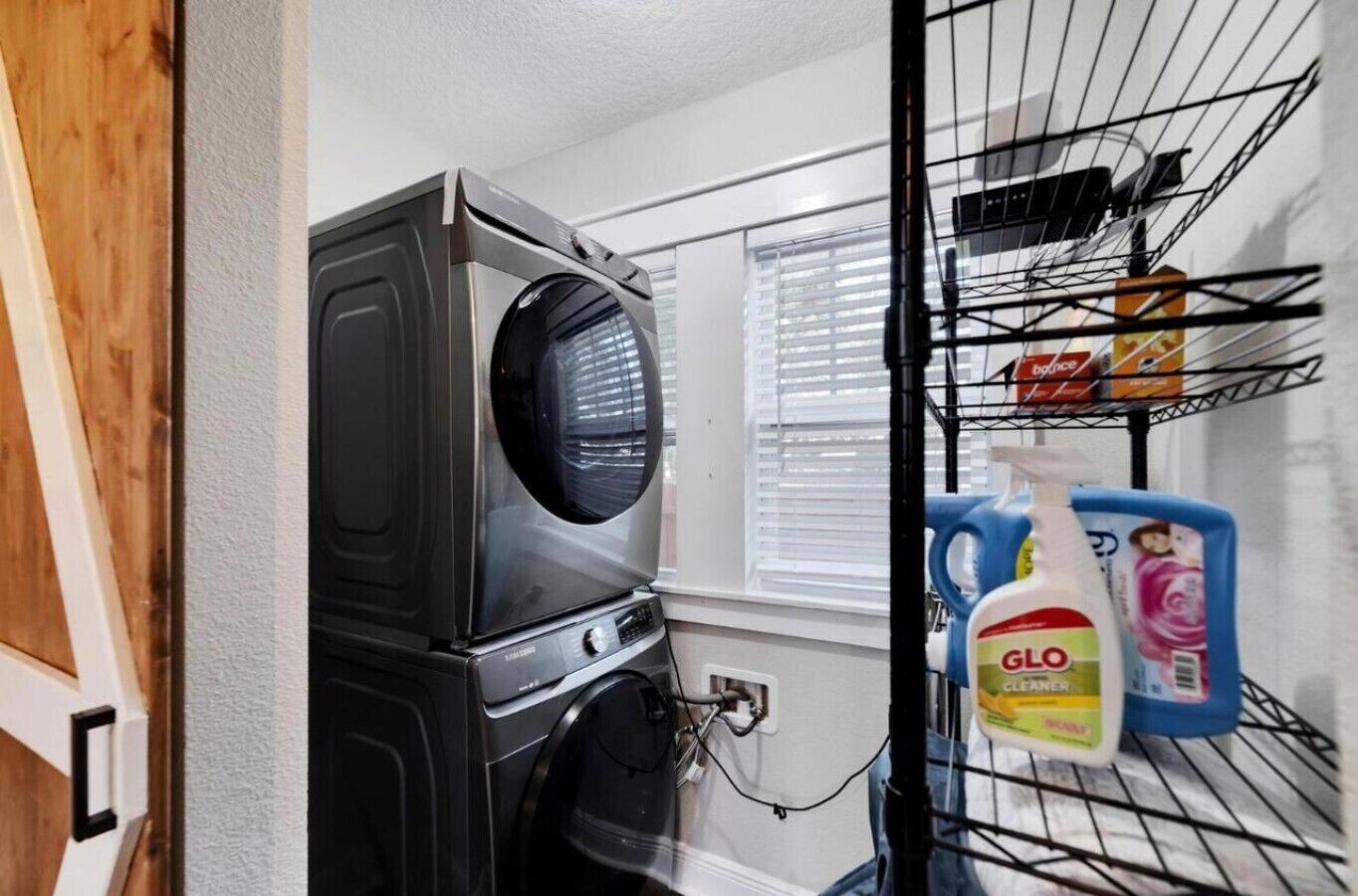 Un espace buanderie comprenant un lave-linge, une étagère avec des produits de nettoyage et une fenêtre avec des stores.