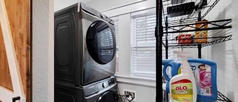 Un espace buanderie comprenant un lave-linge, une étagère avec des produits de nettoyage et une fenêtre avec des stores.