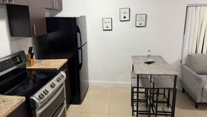 Cocina compacta con estufa, horno y refrigerador, una pequeña mesa de comedor con bancos, y un sofá gris.