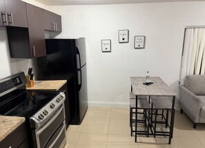 A compact kitchen with a stove, oven, and refrigerator, a small dining table with benches, and a grey sofa. - Miami Central Rest Pad for 2 (Miami)