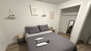 Un dormitorio con cama, almohadas, una mesita de noche, un pequeño estante con plantas y decoración en la pared.