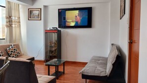 Een woonkamer met een bank, een glazen salontafel, een aan de muur bevestigde televisie en een deur.
